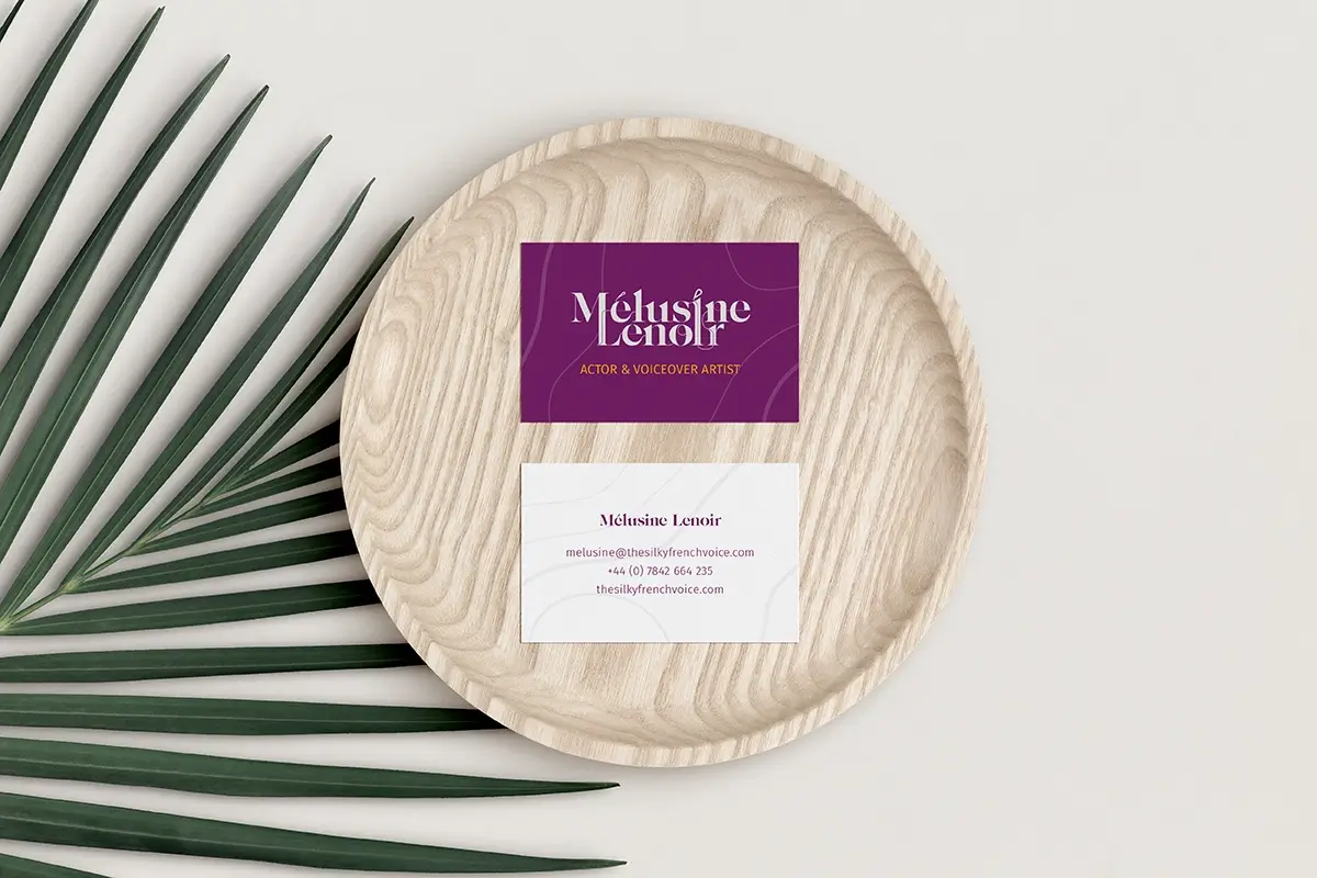 Melusine Lenoir's Actor & Voiceover Artist business card on a pale wooden plate with a palm leaf behind.